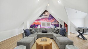 Una sala de estar moderna con un sofá seccional gris, una mesa de centro de madera y una mesa de fútbol. Un mural de un paisaje urbano adorna el techo inclinado.
