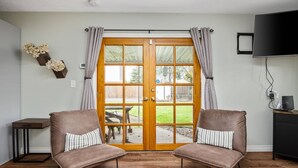 A room with a sliding glass door leading to an outdoor area, two brown cushioned chairs, a small table, a TV on a stand, and a wall-mounted shelf with decorative items. - Splendid Location, Single Level, Pet Friendly, Huge Fenced Yard, BBQ, Games, Next to School Fields (Hillsboro)