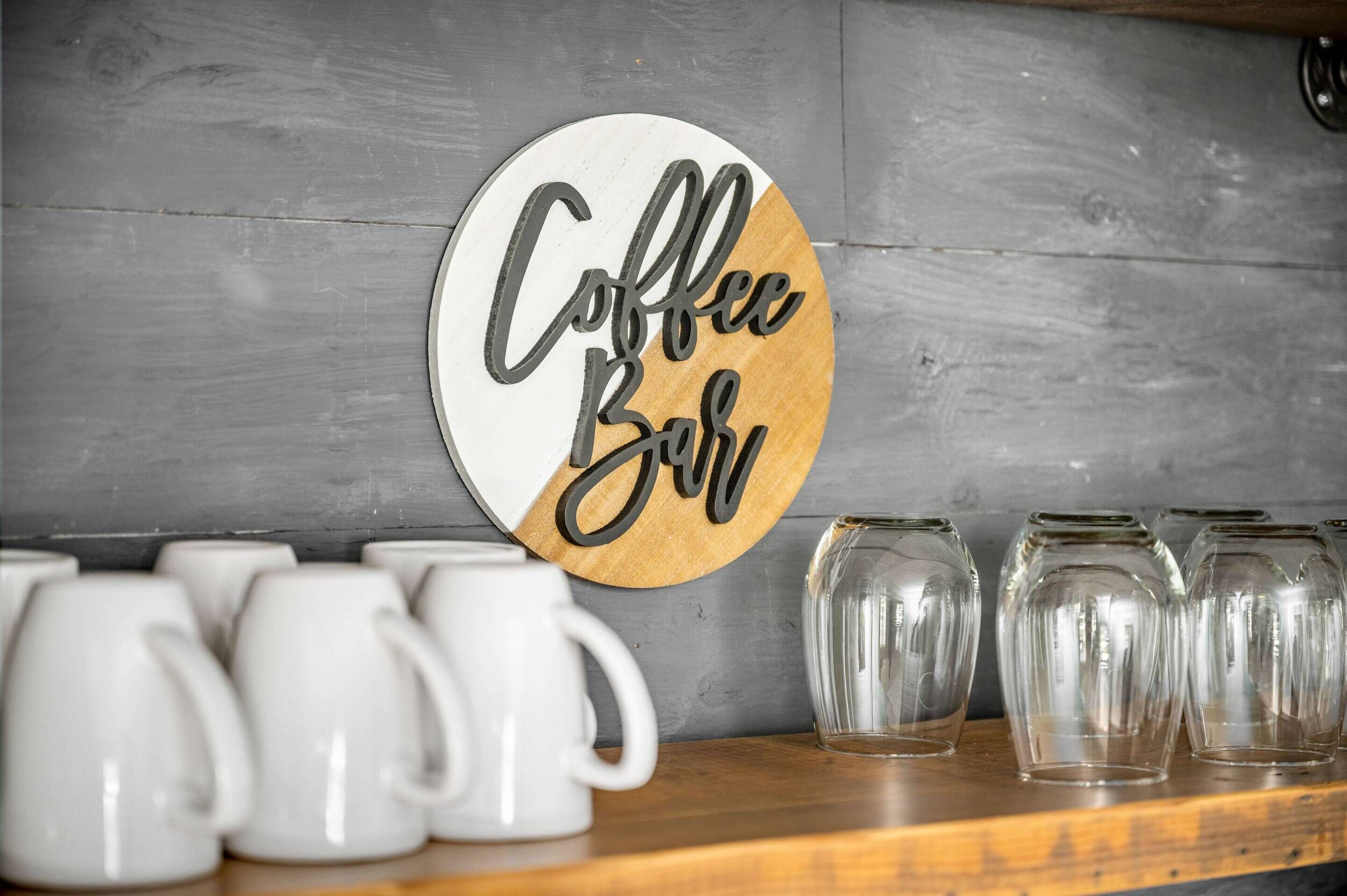 A wall with a circular sign that reads "Coffee Bar" and several white mugs and clear glass cups on a wooden shelf.