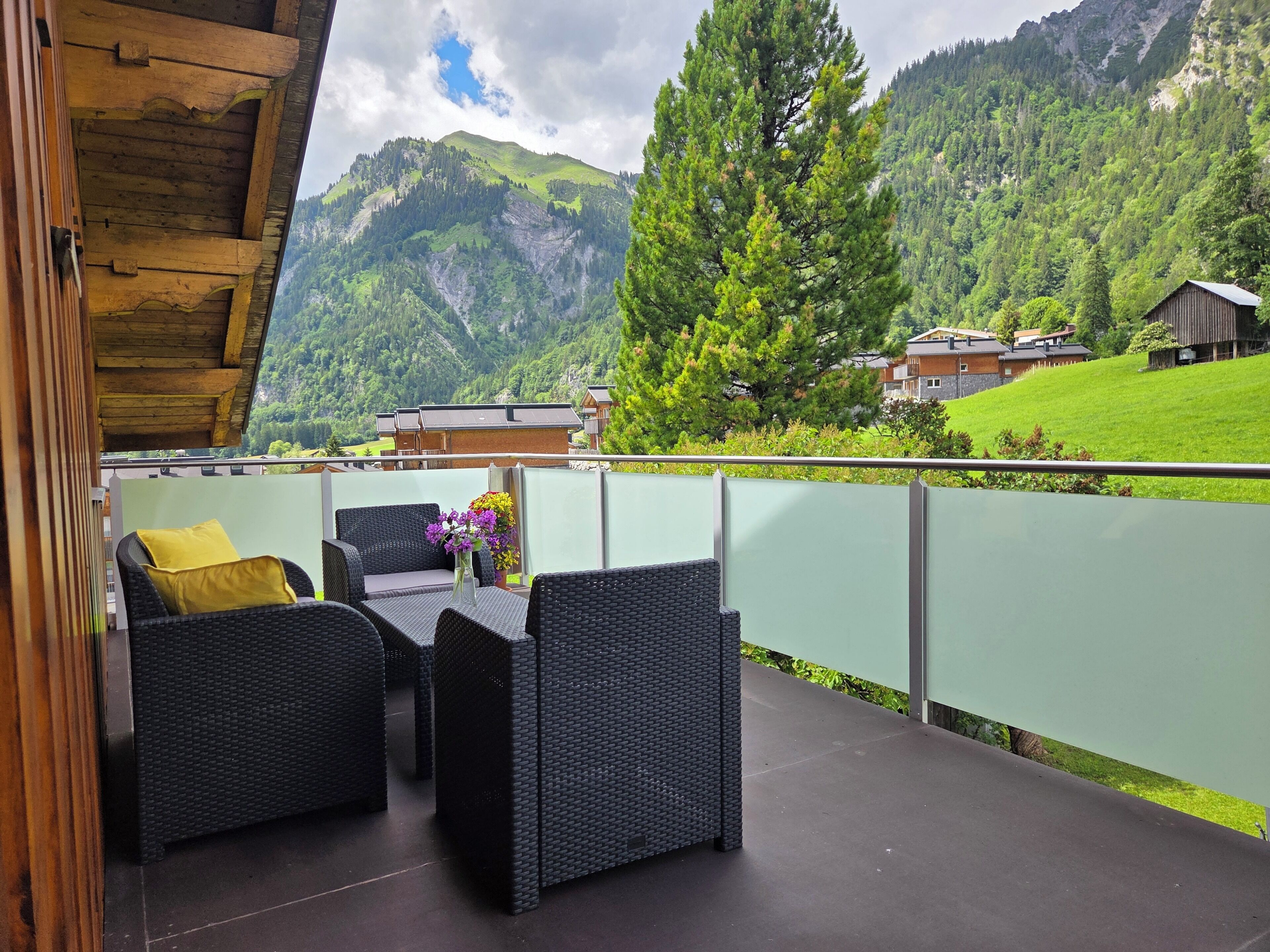 An outdoor seating area with wicker furniture and a vase of flowers, overlooking a mountainous landscape with houses in the background.