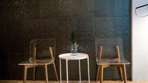 Dos sillas transparentes con una mesa blanca delante, un jarrón pequeño con una planta, y una pared con textura al fondo.