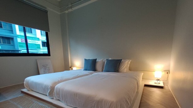 Una cama doble con ropa de cama blanca y almohadas azules, una mesita de noche con lámpara, y una ventana con vistas a edificios.