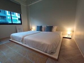 Una cama doble con ropa de cama blanca y almohadas azules, una mesita de noche con lámpara, y una ventana con vistas a edificios.