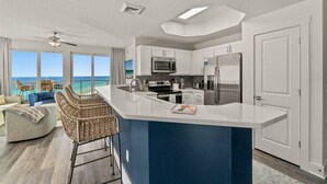 Una cocina moderna con una isla grande, muebles blancos y electrodomésticos de acero inoxidable. Hay un balcón con vista al océano.
