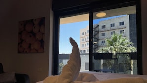 Een bed met een witte deken, een schilderij van sinaasappels aan de muur, en een uitzicht op gebouwen en een palmboom door een raam.