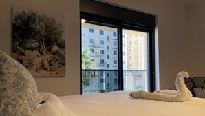 Een slaapkamer met een groot bed, een schilderij van bomen, een raam met uitzicht op gebouwen en een handdoek in de vorm van een zwaan op het bed.