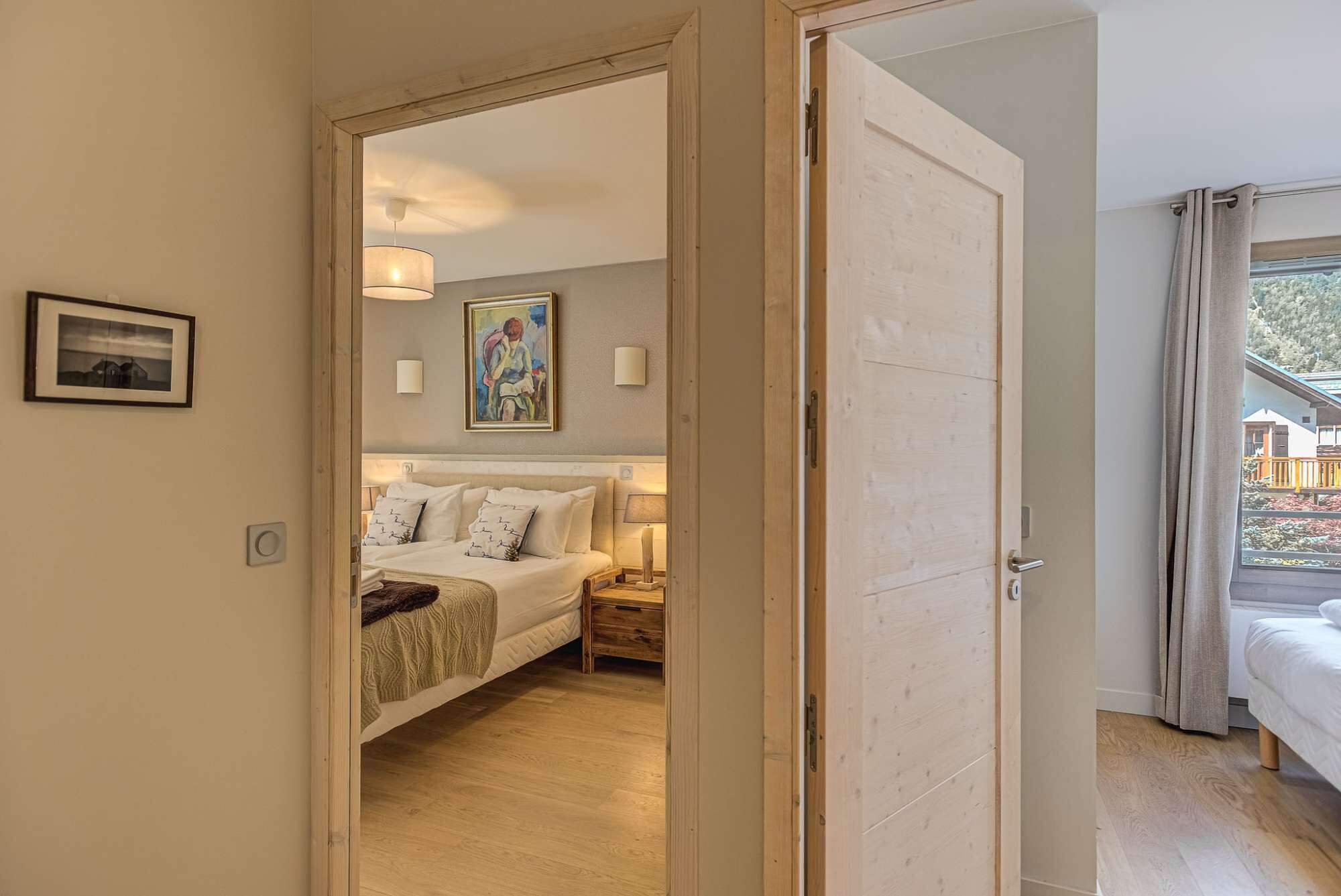 A bedroom with a bed, bedside tables, a painting, and a view through an open door.