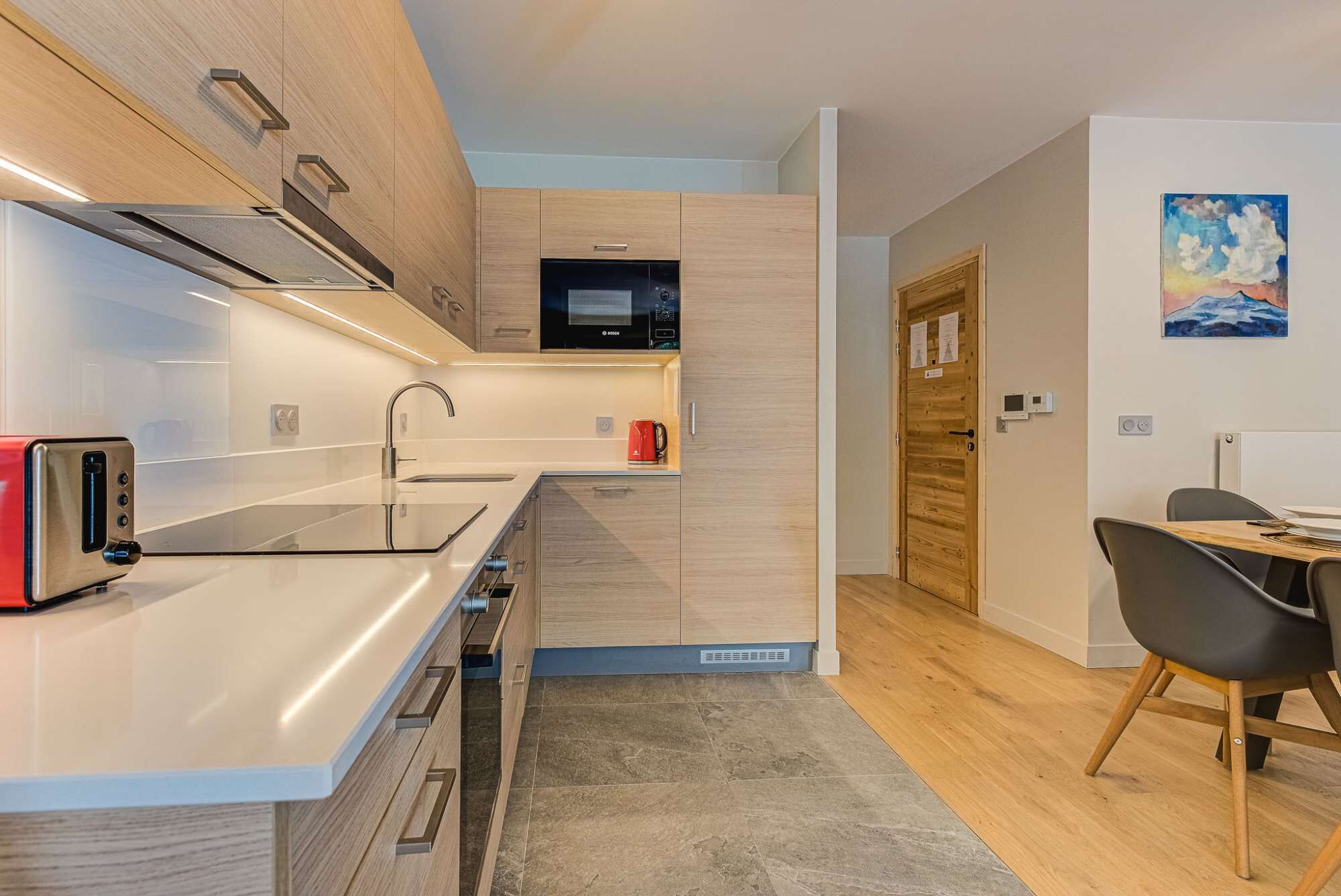 A modern kitchen with a built-in microwave, a red kettle, and a toaster. There is a wooden door and a painting on the wall.