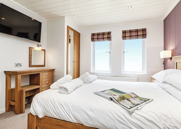 A bedroom with a bed, a TV, a desk, a mirror, and two windows with curtains.