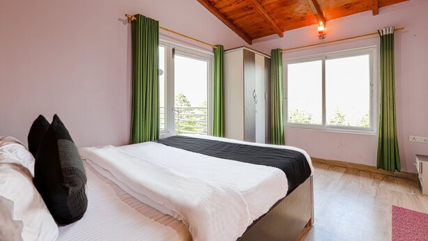 A bedroom with a wooden ceiling, two windows with green curtains, a bed with white and black bedding, and a small bedside table.