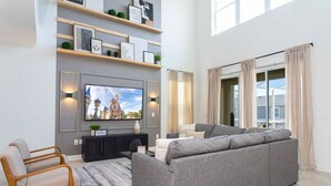 Een moderne woonkamer met een grijze bank, houten stoelen, een flatscreen televisie en een wandplank met decoratieve objecten.