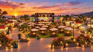 An outdoor dining area with tables and umbrellas, a stage with a colorful backdrop, and a sunset in the background.