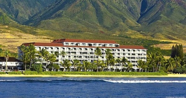 Un hôtel à plusieurs étages, avec un toit rouge, situé près d’une plage bordée de palmiers et avec des montagnes en arrière-plan.