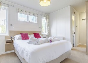 A neatly made bed with white linens and pink pillows, a bedside table with a lamp, and a window with a curtain.