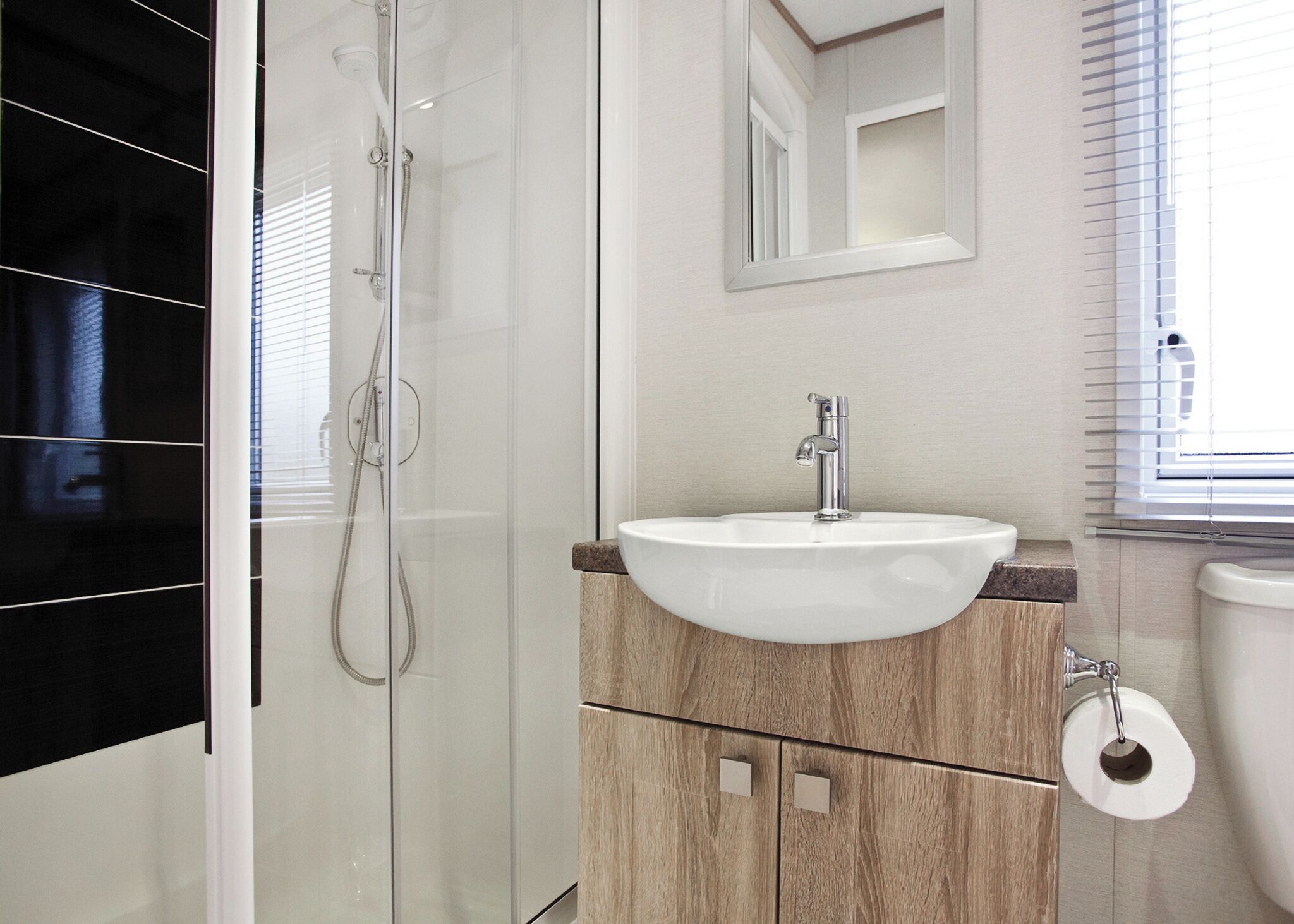 A bathroom with a glass shower enclosure, a white sink with a wooden vanity, and a toilet with a paper towel holder.