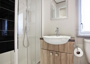 Een badkamer met een douchecabine van glas, een witte wastafel met een houten kastje en een toilet met een handdoekhouder voor keukenpapier.