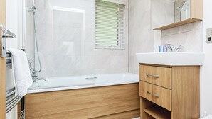 A modern bathroom with a freestanding bathtub, a wooden vanity, a mirror, and a window with blinds. - 1 bedroom accommodation in Newton Saint Loe, Nr Bath (Newton Saint Loe, Nr Bath)