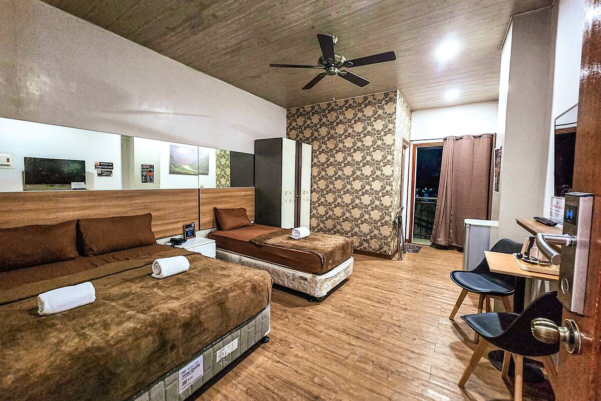 Une chambre d’hôtel avec un plancher en bois, deux lits avec une literie marron, un bureau avec une chaise, un ventilateur de plafond et une porte donnant sur un balcon.