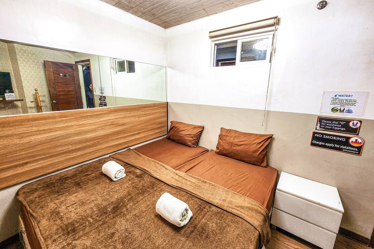 Une chambre d’hôtel avec un lit, une petite table de chevet, un miroir et une fenêtre. Des panneaux sont affichés sur le mur pour rappeler de faire attention à la consommation d’eau et interdisant de fumer.
