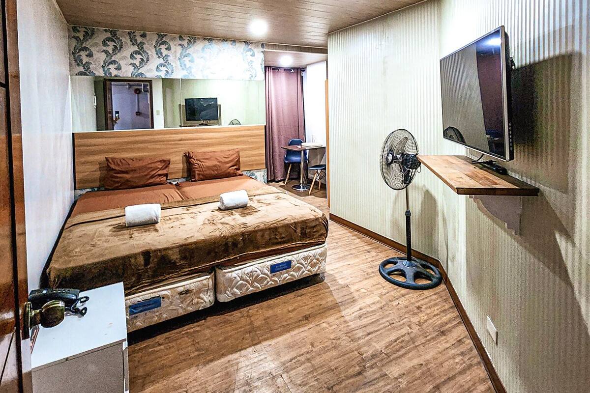 Une pièce de taille modeste avec un plancher en bois, un lit avec un matelas et des oreillers, un petit bureau avec une chaise, un téléviseur fixé au mur et un ventilateur sur pied.