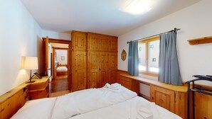 Een slaapkamer met een houten kledingkast, een bed, een bureau met een lamp, en een raam met gordijnen.