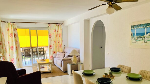 Una sala de estar moderna con sofá, mesa de comedor y sillas. Hay un ventilador de techo, una lámpara y un cuadro en la pared.