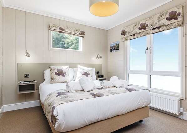 Une chambre à coucher avec un grand lit, une table de chevet, une commode, une fenêtre avec des rideaux et une vue sur l’extérieur.