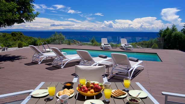 Ein Esstisch mit Frühstücksgegenständen, ein Pool und Liegestühle auf einer Terrasse mit Meerblick.