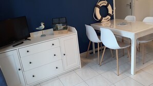 Une pièce avec un meuble blanc, un téléviseur, une table avec deux chaises blanches, une horloge et un gilet de sauvetage accroché au mur.
