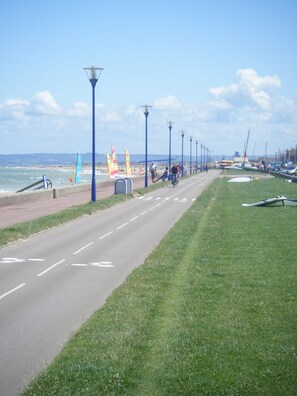 A coastal road with a grassy median, street lamps, and a view of the beach with flags and boats. - Appartement à 50 m de la mer (Ouistreham)