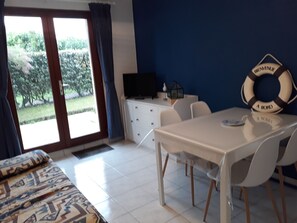 A room with a bed, a dining table with chairs, a TV on a cabinet, and a view of greenery outside. - Appartement à 50 m de la mer (Ouistreham)