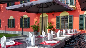 A dining table set with white plates, silverware, and wine glasses, surrounded by black chairs, under a large umbrella, against a building with green shutters and potted plants. - Villa in Piedmont with private pool in beautiful vineyard of Tenuta CostaRossa (Piemonte)