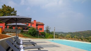 A poolside area with lounge chairs and umbrellas, a red building in the background, and a clear sky. - Villa in Piedmont with private pool in beautiful vineyard of Tenuta CostaRossa (Piemonte)