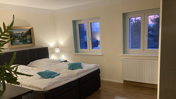 Ein Schlafzimmer mit Bett, Nachttisch, Lampe und einem Fenster mit Blick auf Bäume.