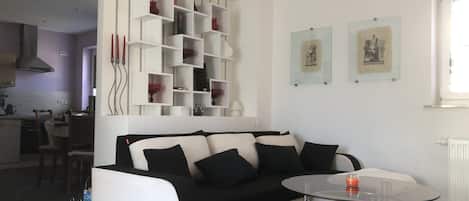 A modern living room with a black and white sofa, a glass coffee table, and a white shelving unit against the wall.