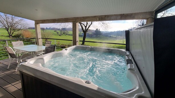 Ein Whirlpool auf einer Holzterrasse mit Tisch und Stühlen, mit Blick auf eine grüne Landschaft.
