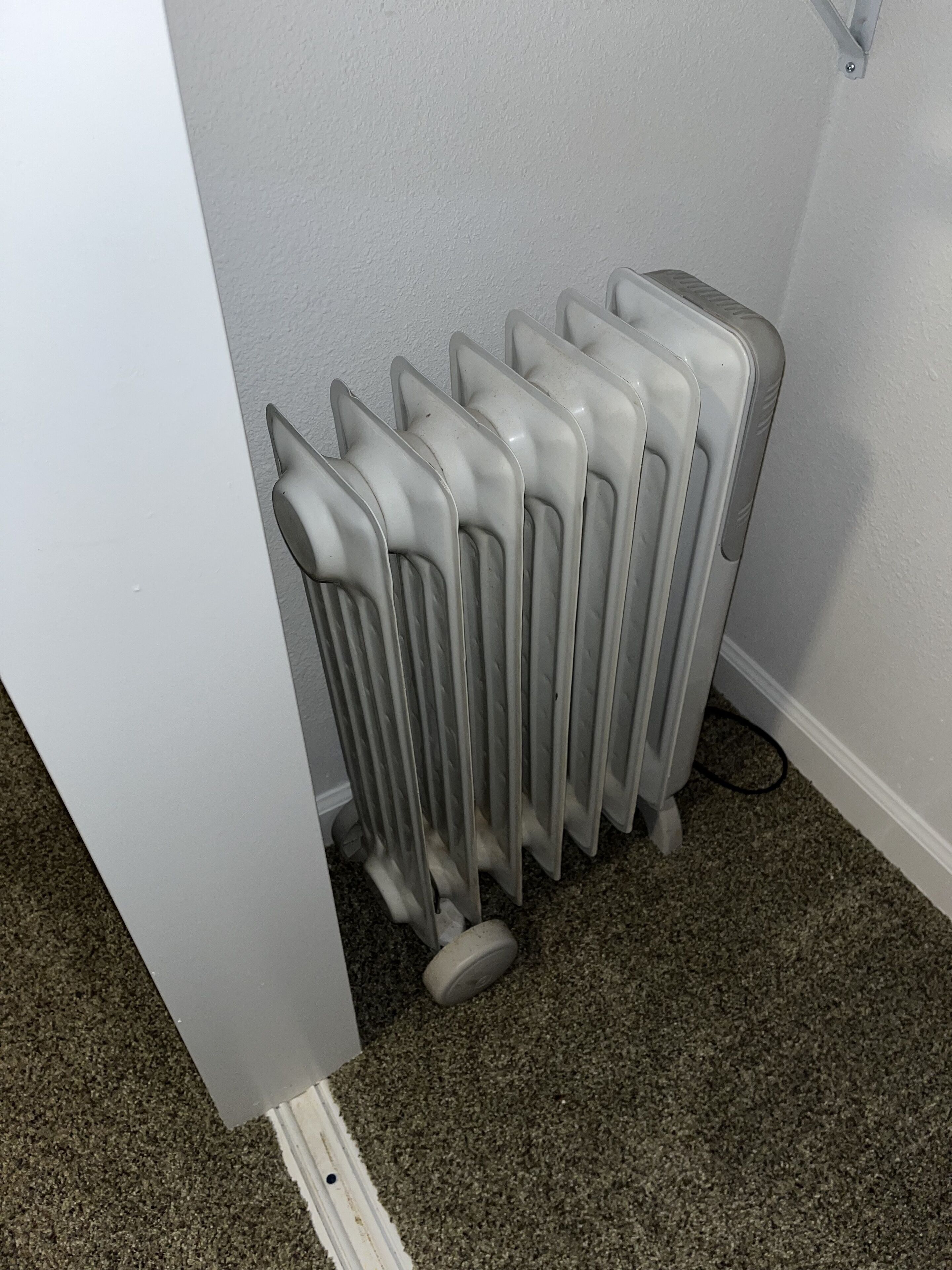 A radiator in a corner with a white wall and carpeted floor.