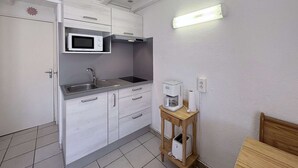 Cocina compacta con muebles blancos, microondas, lavamanos, cafetera y una mesita de madera.