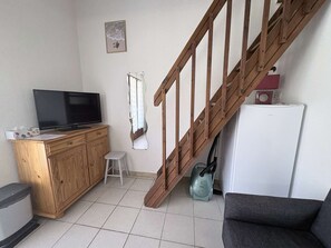 Un espacio habitable pequeño con una escalera de madera, un televisor sobre un mueble de madera, un taburete, una aspiradora y un refrigerador.