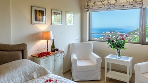 A bedroom with a bed, a bedside table with a lamp, a chair, a small table with a vase of flowers, and a window with a view of the sea. - Luxury Villa Hara with private pool (Malevizi)