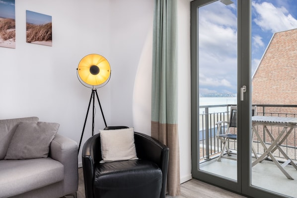 A modern living room with a sofa, a black leather chair, a floor lamp, and a view of the balcony and cityscape through a glass door.