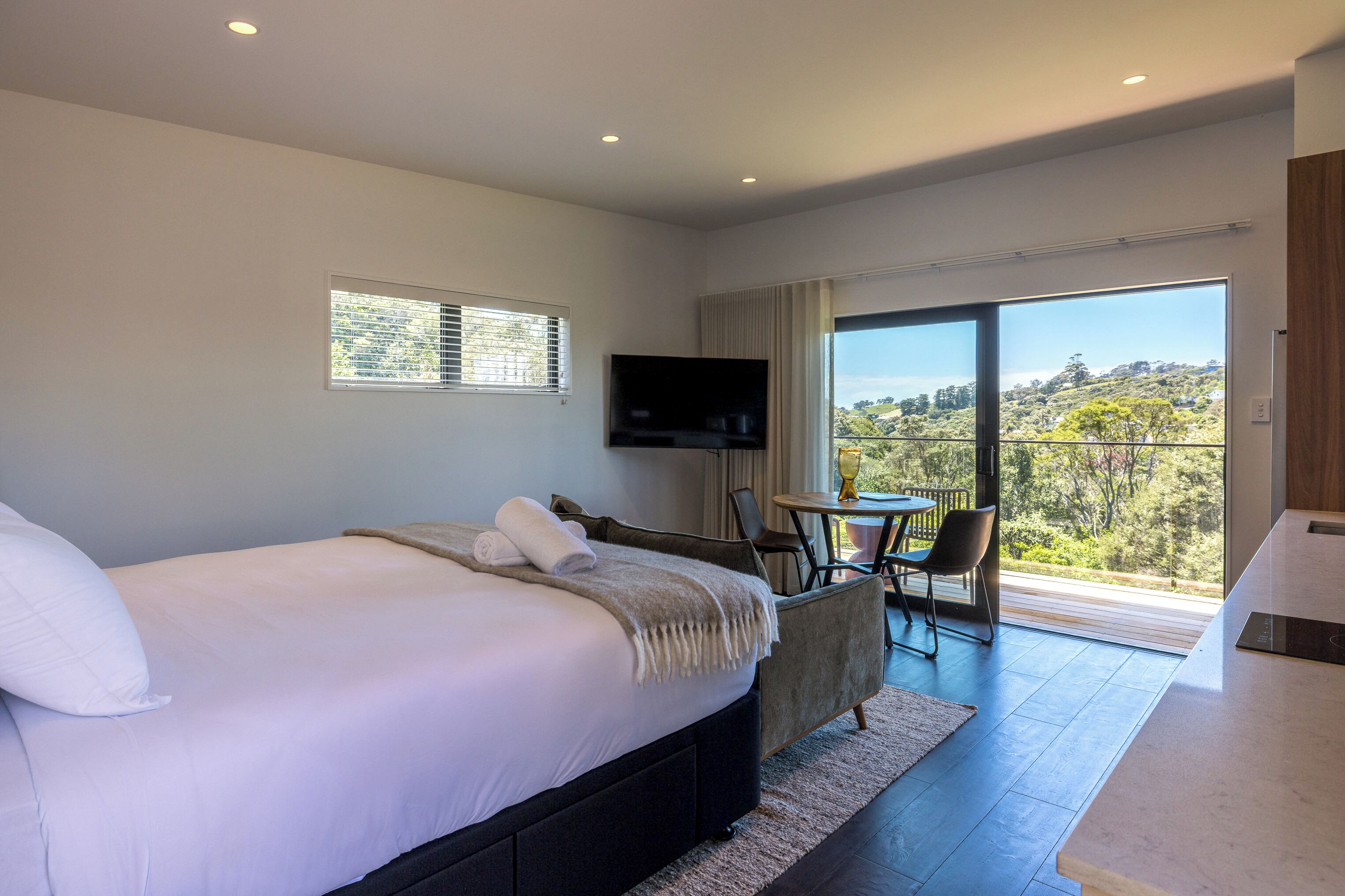 A modern bedroom with a large bed, a TV mounted on the wall, and a balcony with a view of the outdoors.