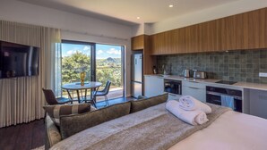 A modern hotel room with a large bed, a dining area with a table and chairs, a kitchen with a large window, and a TV mounted on the wall. - Hidden Valley Waiheke (Waiheke Island)