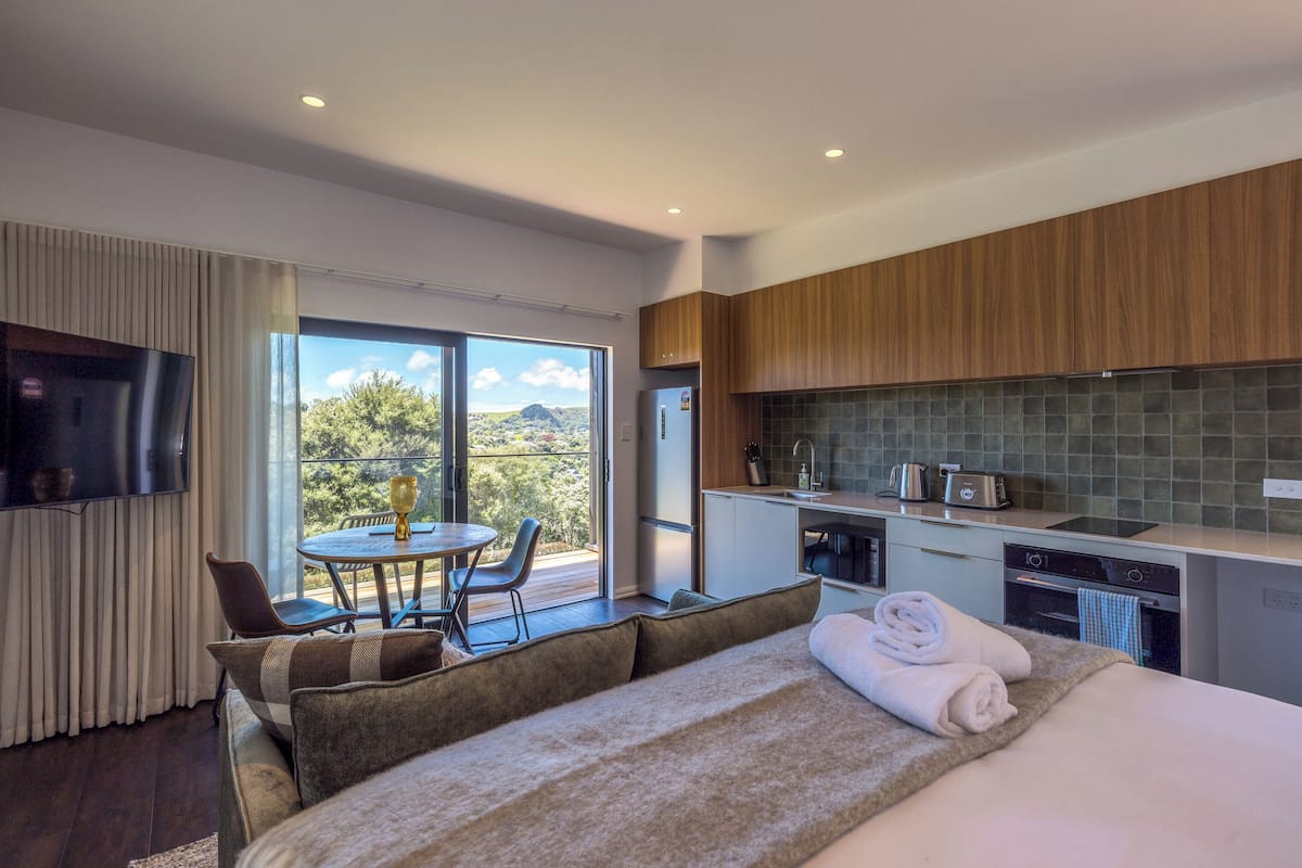 A modern hotel room with a large bed, a dining area with a table and chairs, a kitchen with a large window, and a TV mounted on the wall.