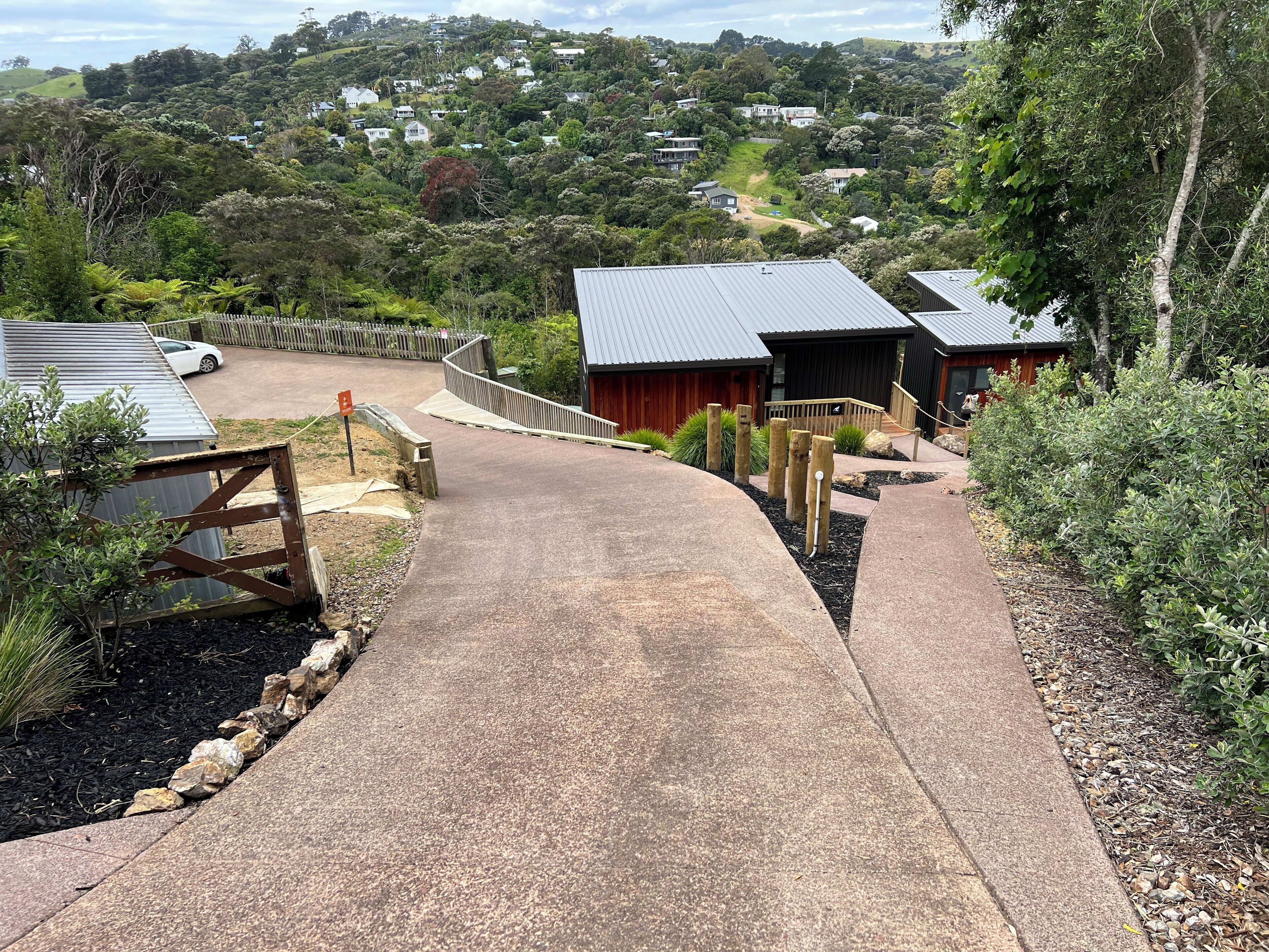 A concrete pathway leading to a building with a metal roof, surrounded by greenery and residential houses in the background.