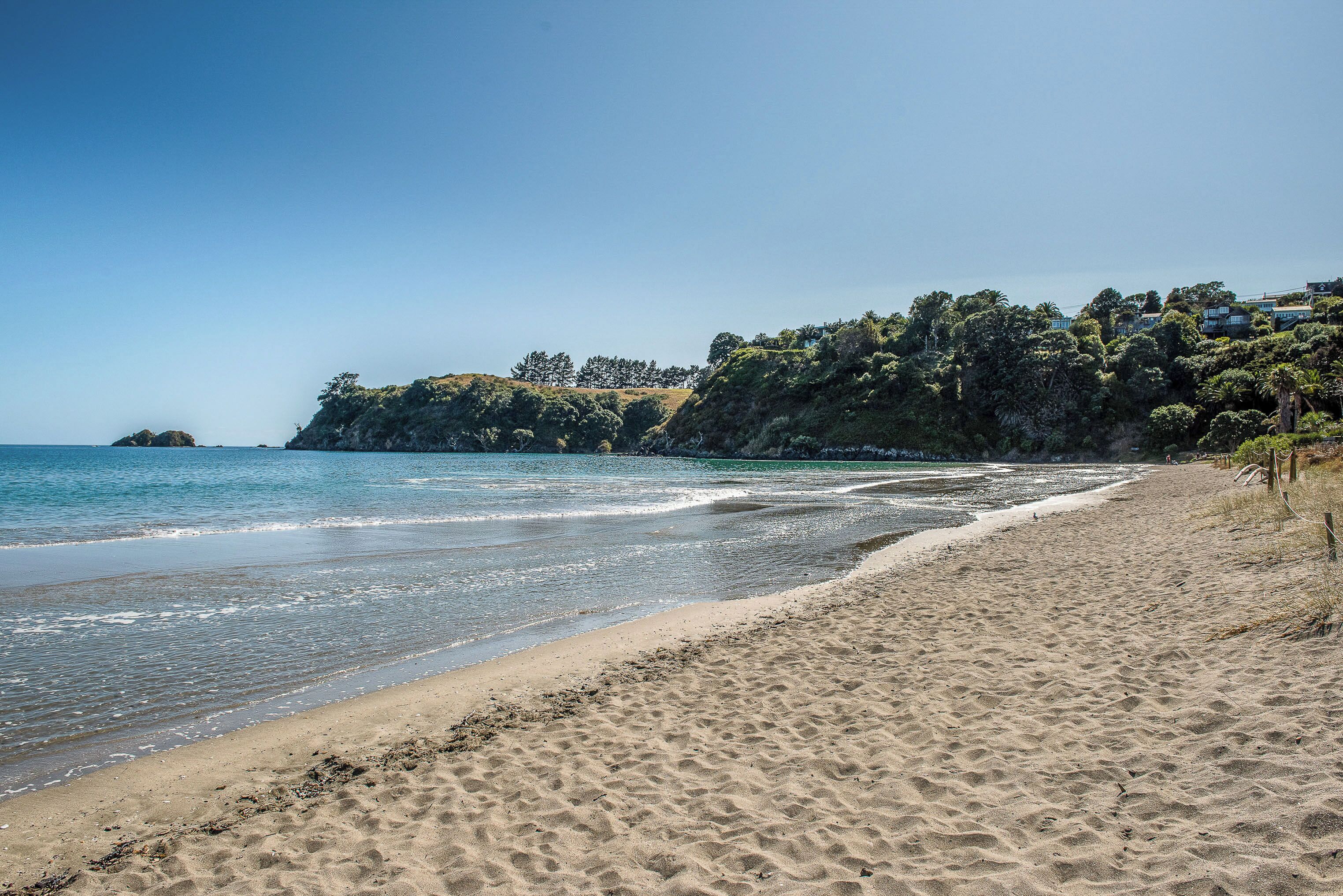 Una playa arenosa con olas suaves, cielo despejado y un acantilado con vegetación y edificios.