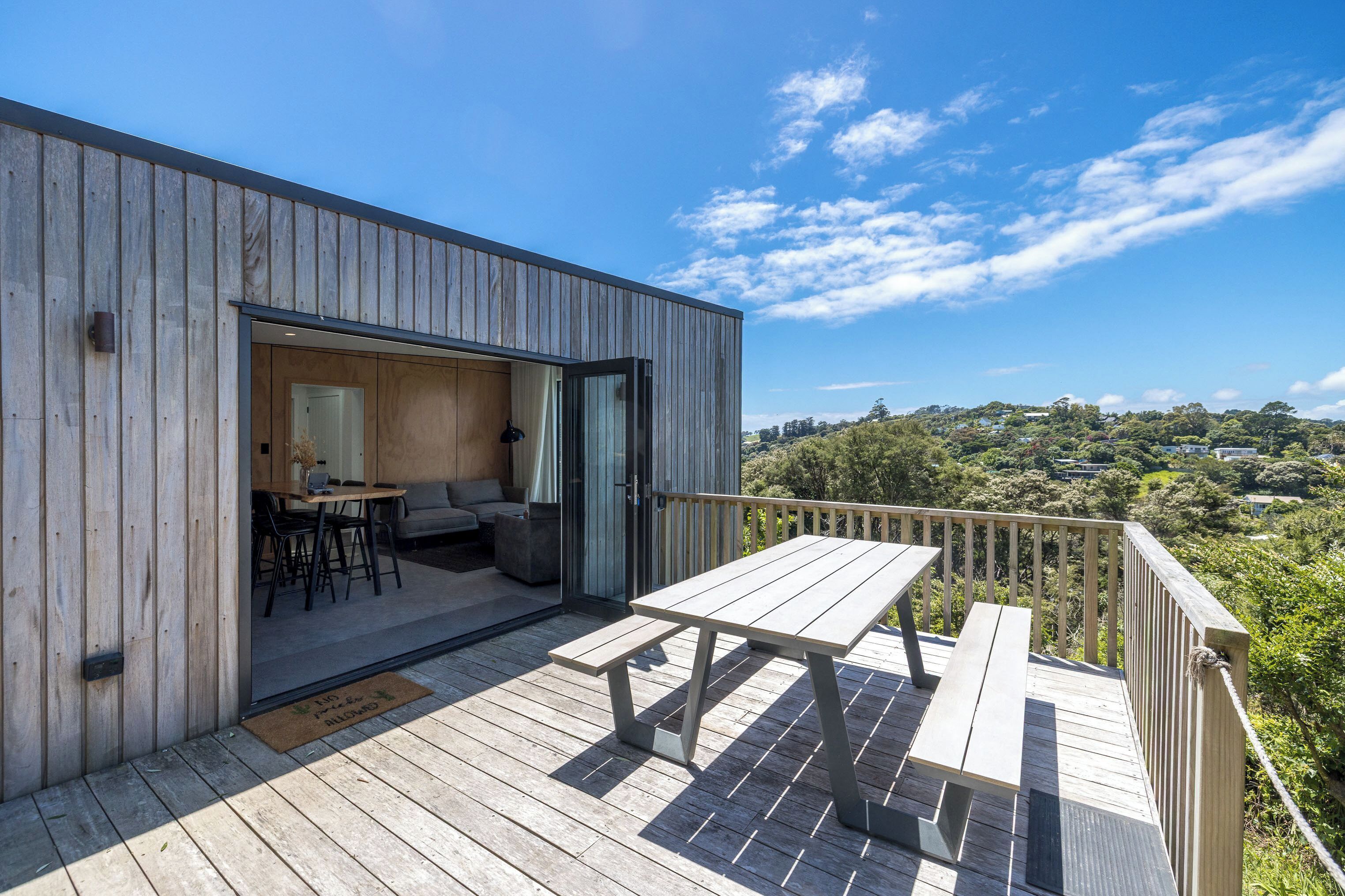 Una terraza de madera con una mesa y bancos, un cerramiento de vidrio corredero que da a un interior con un sofá y sillones, y vistas a un paisaje montañoso.