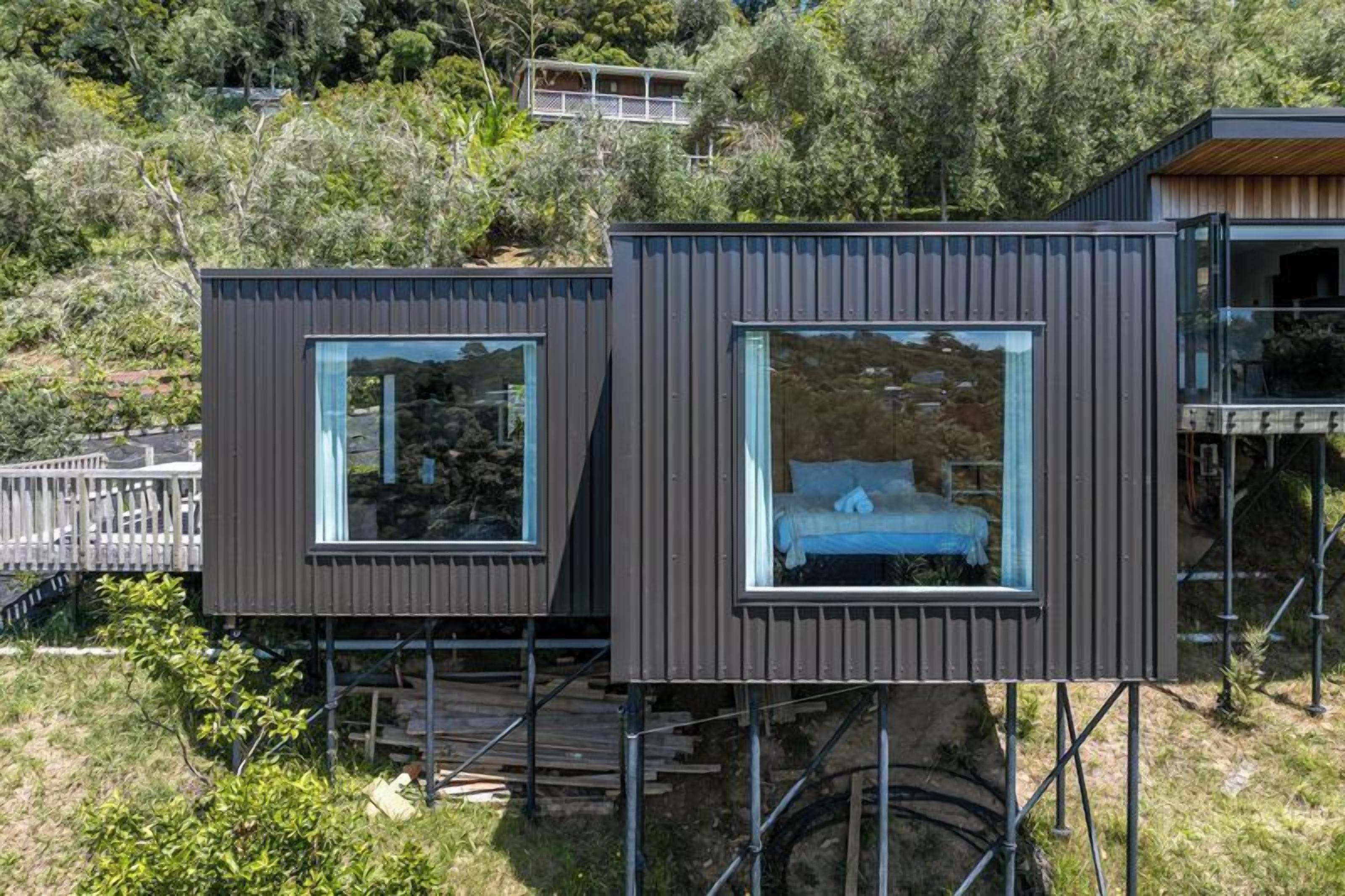 Two modern, elevated wooden cabins with large windows, situated on stilts amidst a forested hillside.