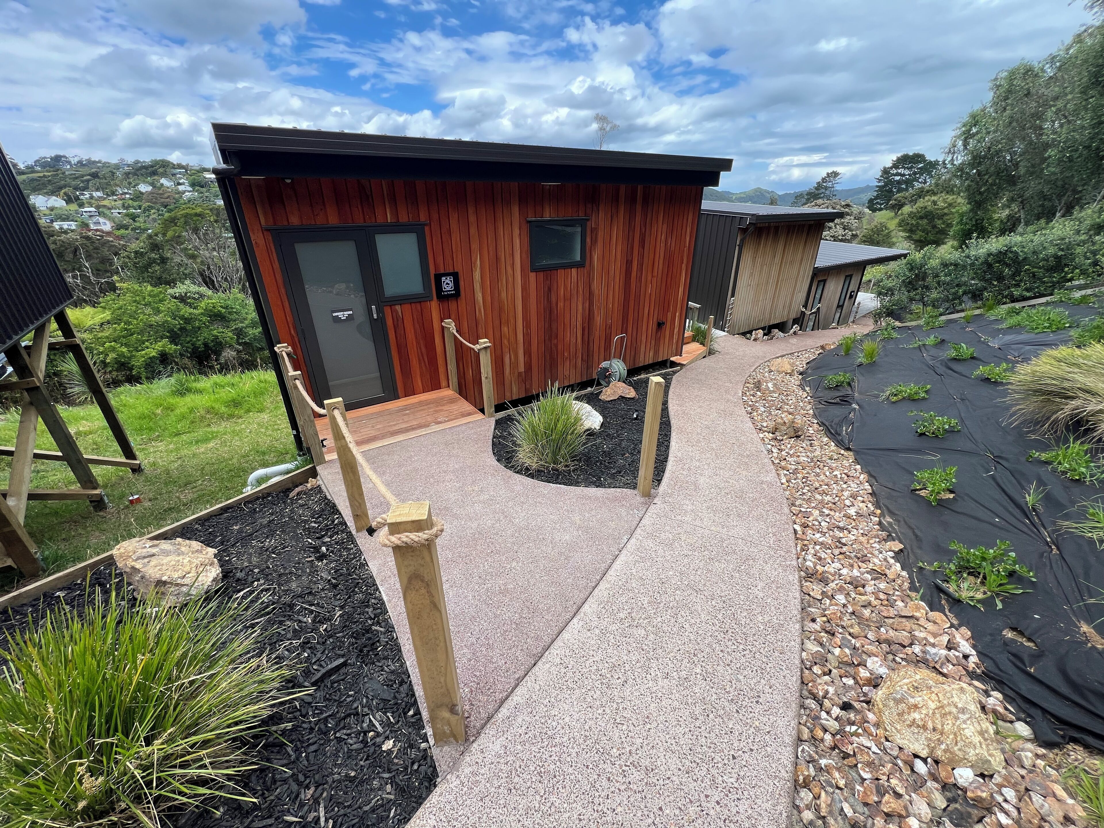 Una cabaña moderna de madera con un sendero de concreto que lleva a la entrada, rodeada de jardines ajardinados y vistas a las colinas.