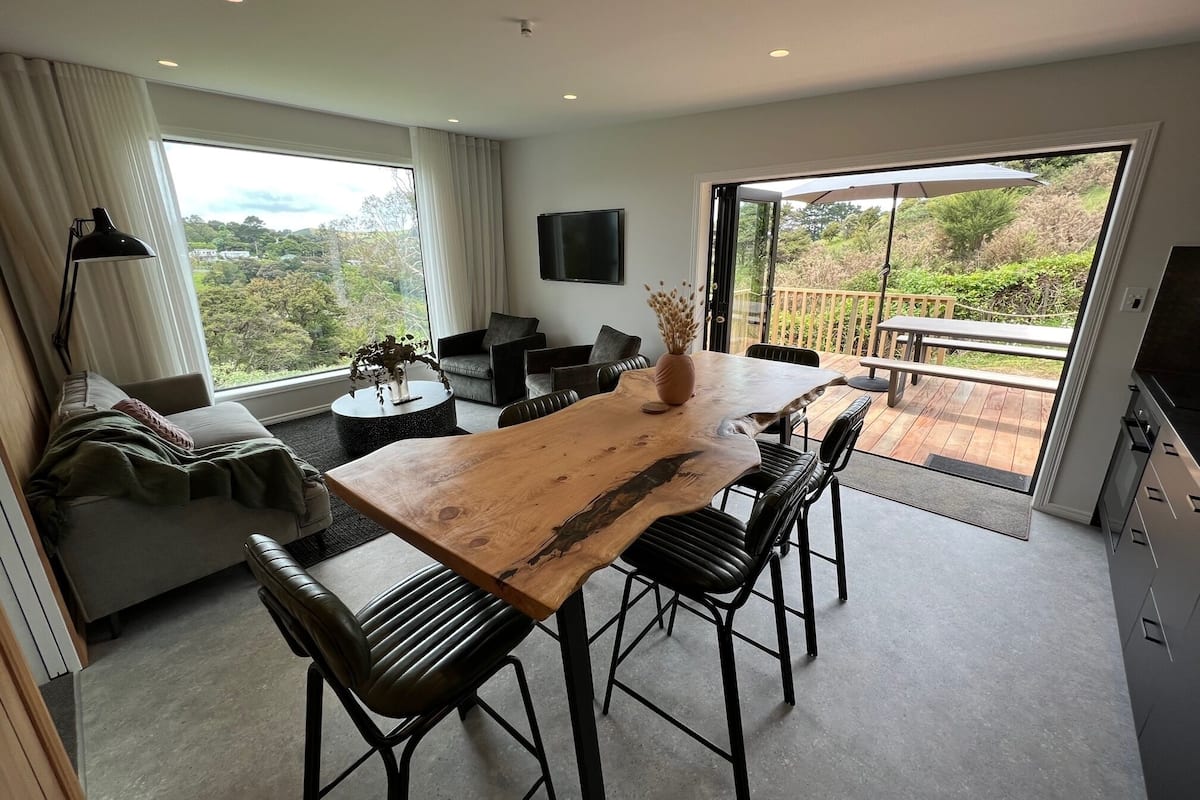 A modern living room with a wooden dining table, black leather chairs, a sofa, and a TV on the wall.
